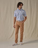 Men's Island Paradise Polo in Cool Blue, full front view on model featuring an all-over tropical print with palm trees, hibiscus flowers, sailboats, and tropical fish and blue on a white body, worn with tan pants, a tan belt, and tan and white golf shoes