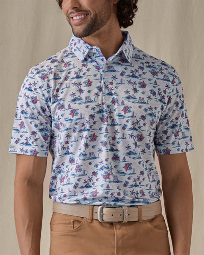 Men's Island Paradise Polo in Cool Blue, front view on model featuring an all-over tropical print with palm trees, hibiscus flowers, sailboats, and tropical fish and blue on a white body, worn with tan pants and a tan belt