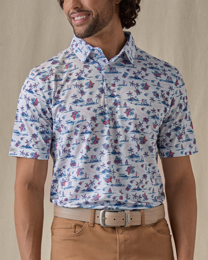 Men's Island Paradise Polo in Cool Blue, front view on model featuring an all-over tropical print with palm trees, hibiscus flowers, sailboats, and tropical fish and blue on a white body, worn with tan pants and a tan belt