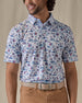 Men's Island Paradise Polo in Cool Blue, front view on model featuring an all-over tropical print with palm trees, hibiscus flowers, sailboats, and tropical fish and blue on a white body, worn with tan pants and a tan belt