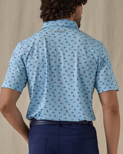 Men's Dog Paddle Polo in Cool Blue, back view on model featuring an all-over print of dogs paddle boarding on red and blue boards on a light blue body with the Straight Down logo embroidered at the upper back, worn with navy pants