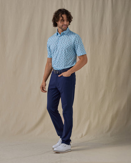Men's Dog Paddle Polo in Cool Blue, full front view on model featuring an all-over print of dogs paddle boarding on red and blue boards on a light blue body, worn with navy pants, a navy belt, and white golf shoes