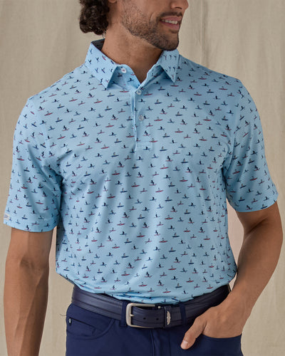 Men's Dog Paddle Polo in Cool Blue, front view on model featuring an all-over print of dogs paddle boarding on red and blue boards on a light blue body, worn with navy pants and a navy belt