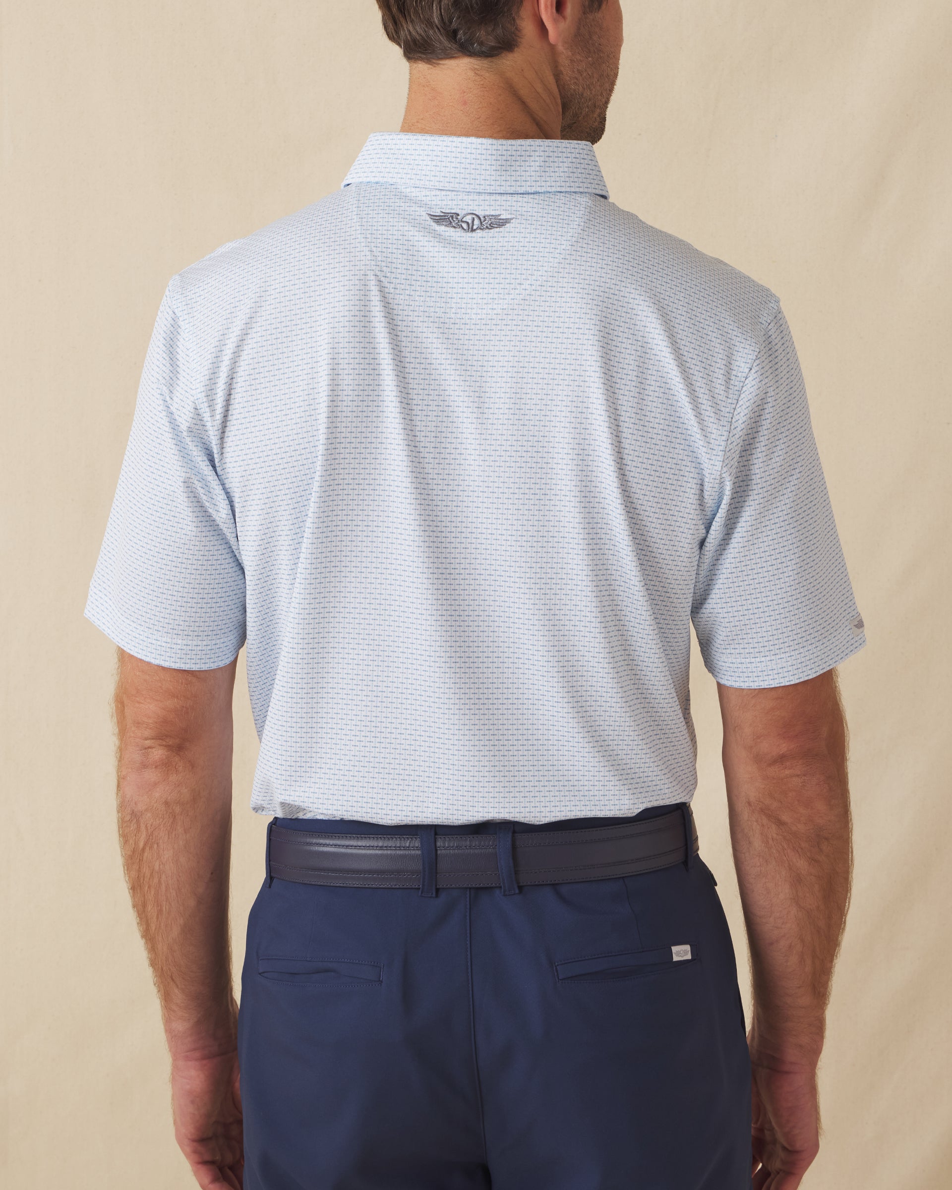 Men's Wilshire Polo in Gemstone, back view on model featuring a white base with an all-over blue geometric micro print, with Straight Down logo embroidered at upper back, paired with navy pants