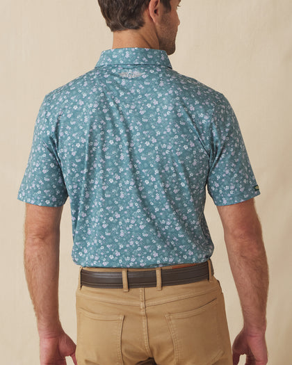 Men's Dogwood Polo in Sage, back view on model featuring a sage green base with a scattered white and lavender floral print and Straight Down logo embroidered at upper back, paired with khaki pants