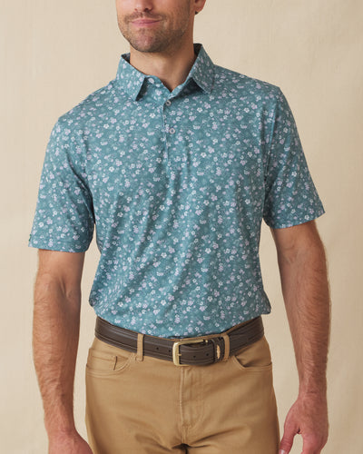 Men's Dogwood Polo in Sage, front view on model featuring a sage green base with a scattered white and lavender floral print, paired with khaki pants and a leather belt