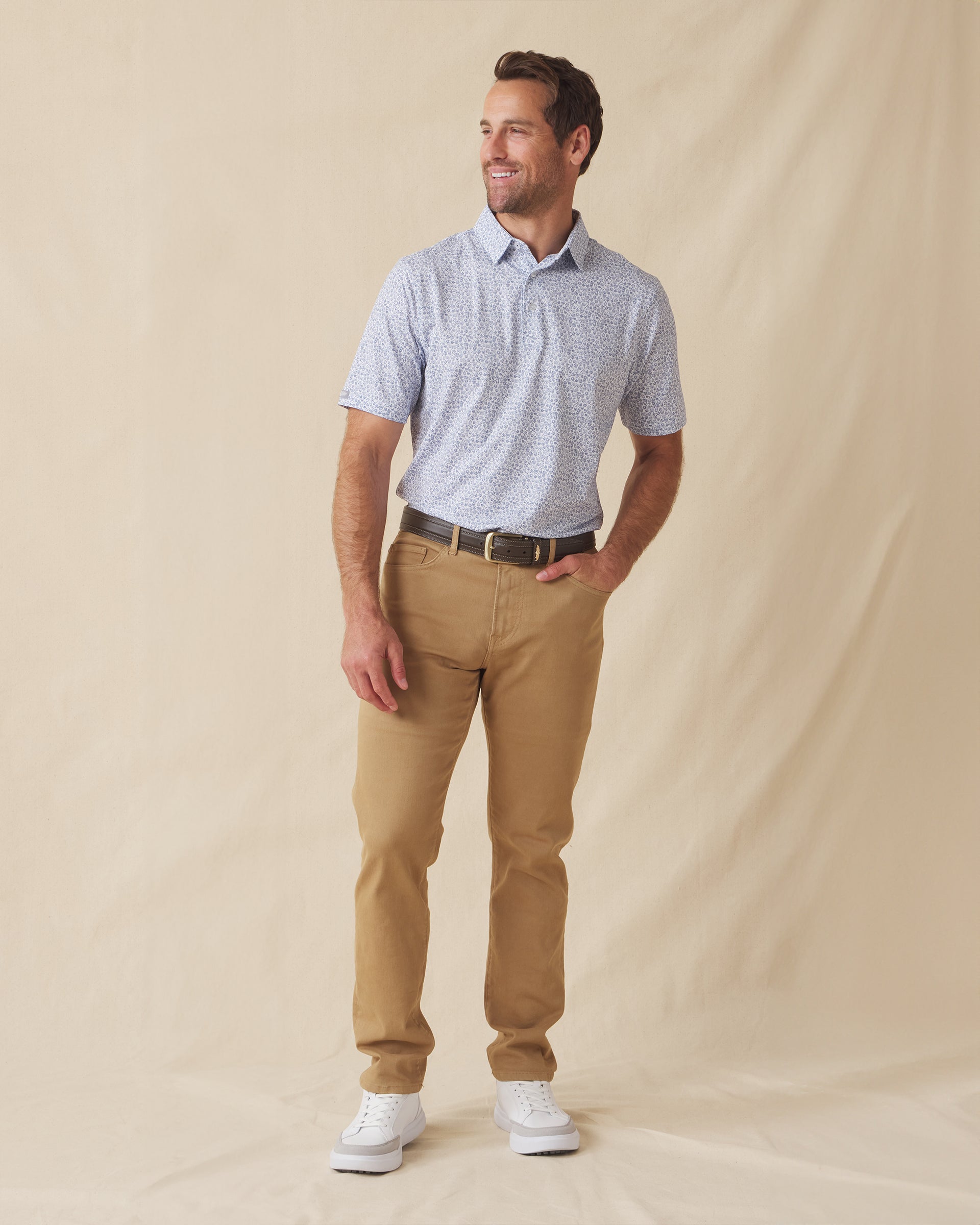 Men's Dog Pound Polo in Moonlight, full length model view featuring a white short sleeve polo with an all-over scattered dog face and bone print in navy paired with khaki pants, a brown belt, and white shoes