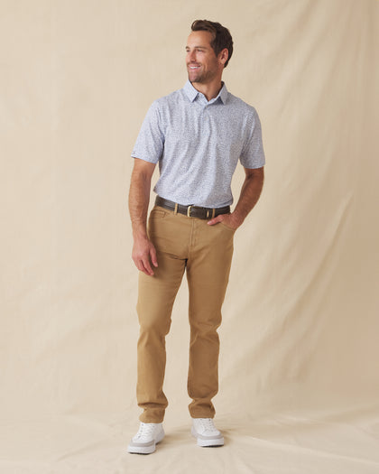 Men's Dog Pound Polo in Moonlight, full length model view featuring a white short sleeve polo with an all-over scattered dog face and bone print in navy paired with khaki pants, a brown belt, and white shoes