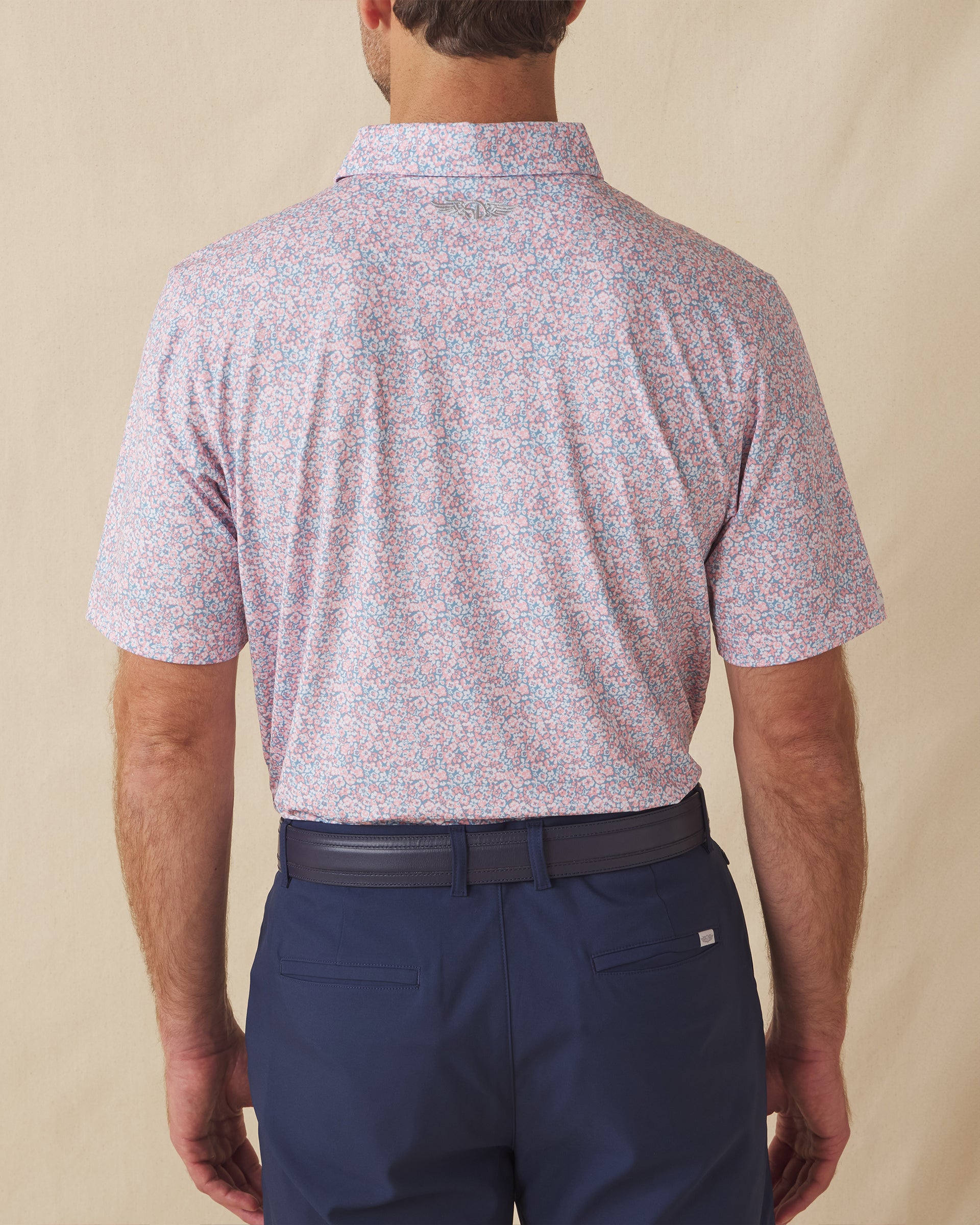 Men's Ocala Polo in Spritz, back view on model featuring a blue base with an all-over pink floral print, with Straight Down logo embroidered at upper back, paired with navy pants