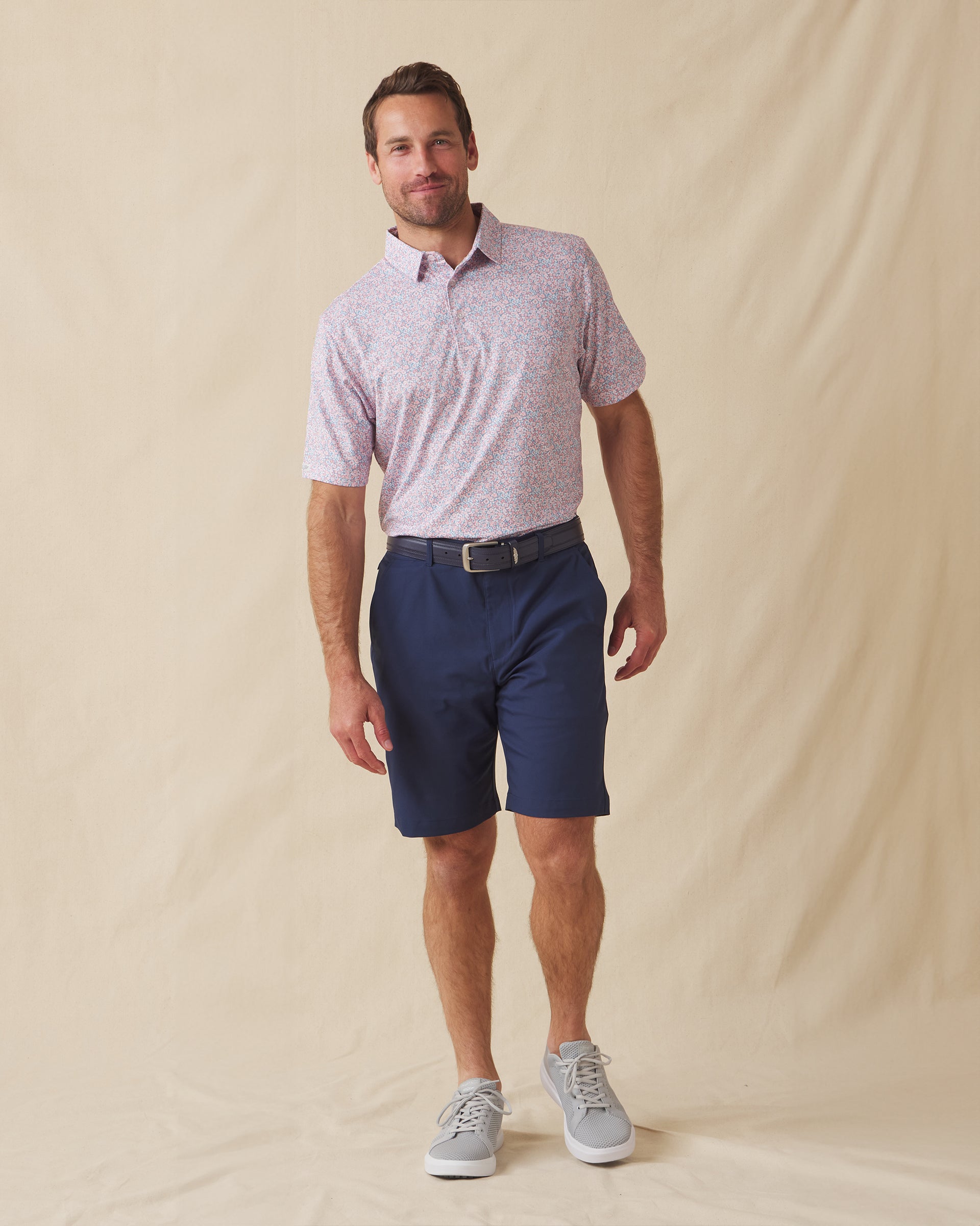 Men's Ocala Polo in Spritz, full length front view on model featuring a blue base with an all-over pink floral print, paired with navy shorts, a dark leather belt, and gray shoes