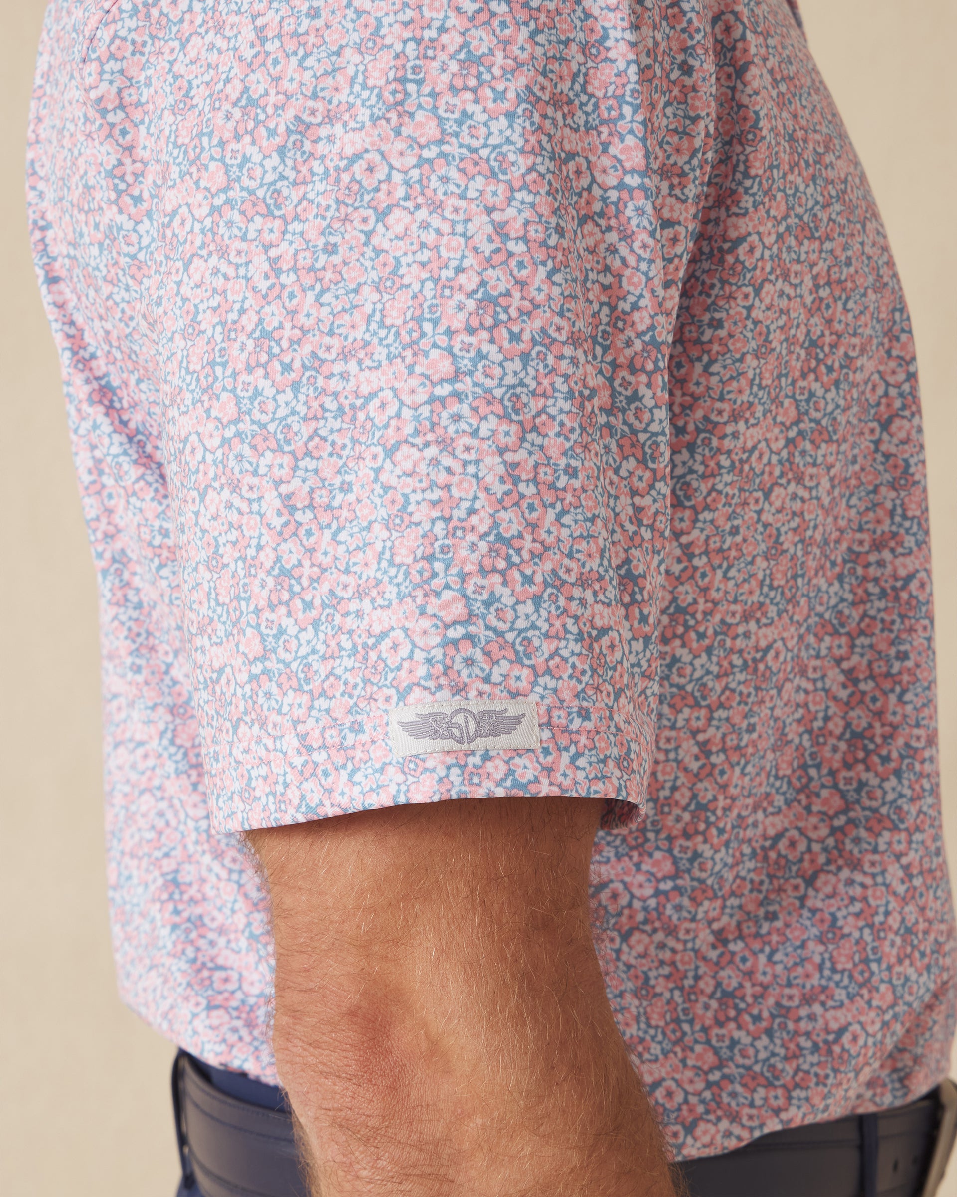 Men's Ocala Polo in Spritz, close-up sleeve view on model highlighting the Straight Down logo patch on the sleeve hem and the all-over pink floral print on a blue base
