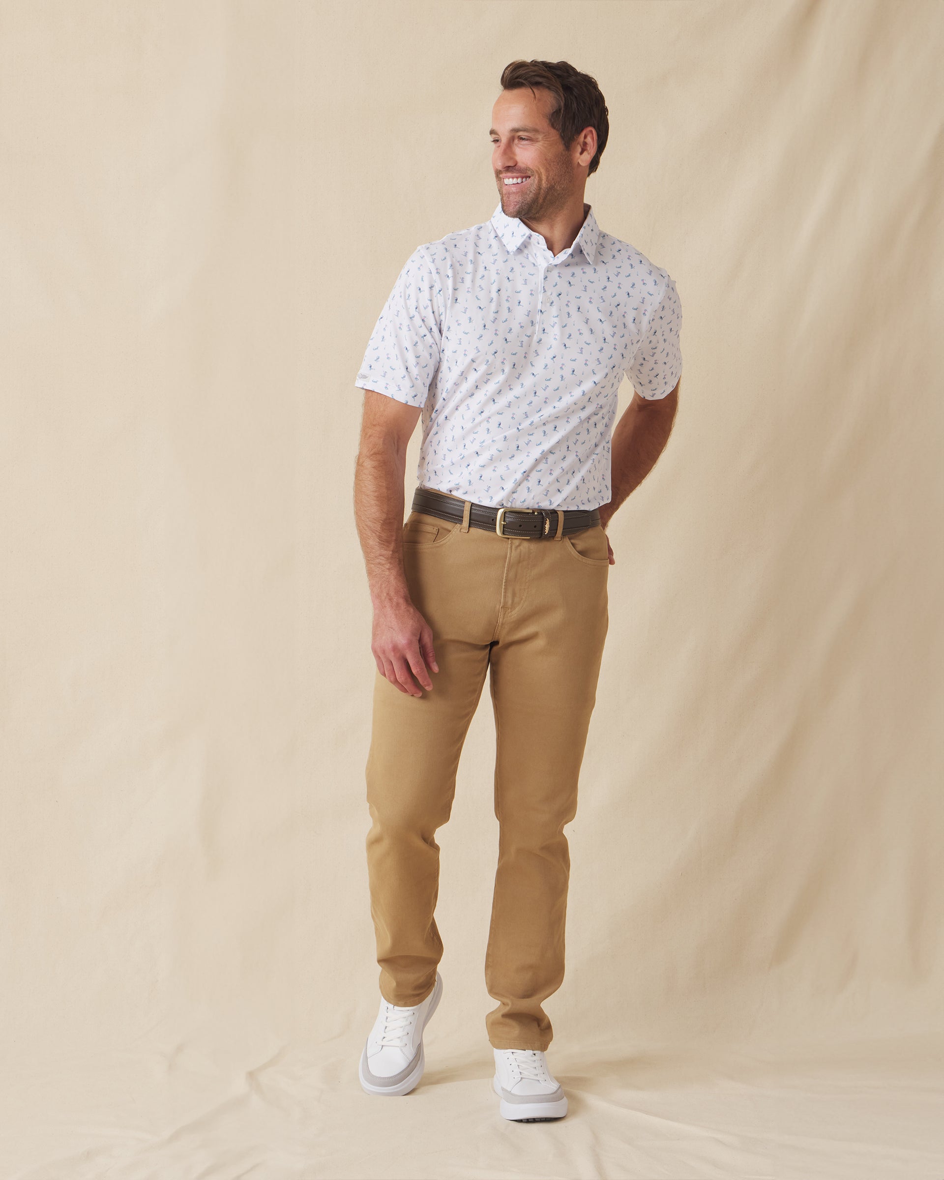 Men's Gators Polo in Violet, full length model view featuring a white short sleeve polo with an all-over scattered golf-playing alligator print in blue and pink paired with khaki pants and white shoes