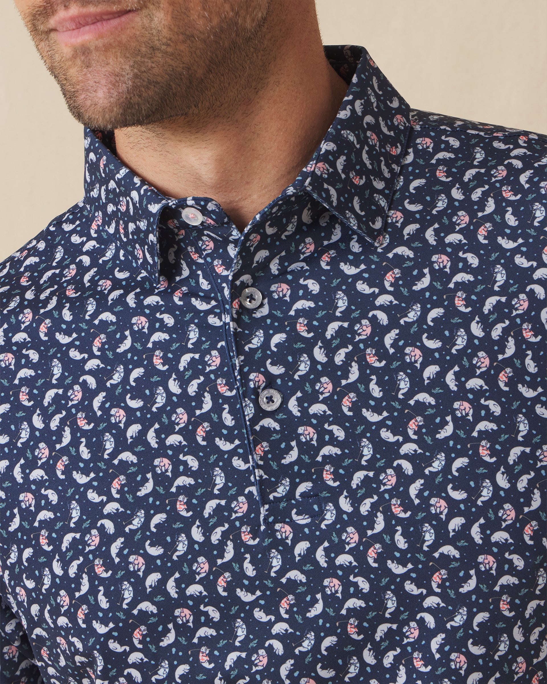Men's Manatee County Polo in New Indigo, close-up model view featuring the collar and button placket detail of a navy short sleeve polo with an all-over scattered golf-playing manatee print in white and pink