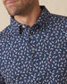 Men's Manatee County Polo in New Indigo, close-up model view featuring the collar and button placket detail of a navy short sleeve polo with an all-over scattered golf-playing manatee print in white and pink