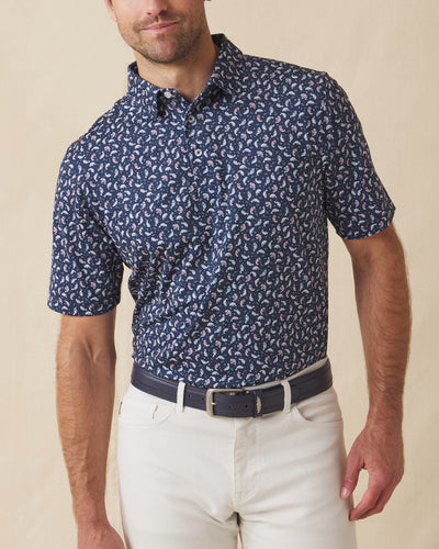 Men's Manatee County Polo in New Indigo, front model view featuring a navy short sleeve polo with an all-over scattered golf-playing manatee print in white and pink paired with white pants and a navy belt