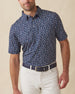 Men's Manatee County Polo in New Indigo, front model view featuring a navy short sleeve polo with an all-over scattered golf-playing manatee print in white and pink paired with white pants and a navy belt