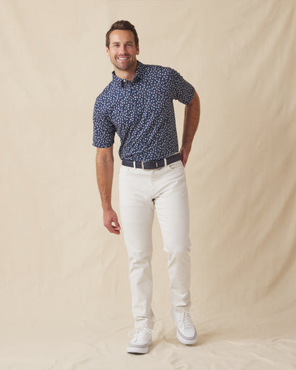 Men's Manatee County Polo in New Indigo, full length model view featuring a navy short sleeve polo with an all-over scattered golf-playing manatee print in white and pink paired with white pants and white shoes