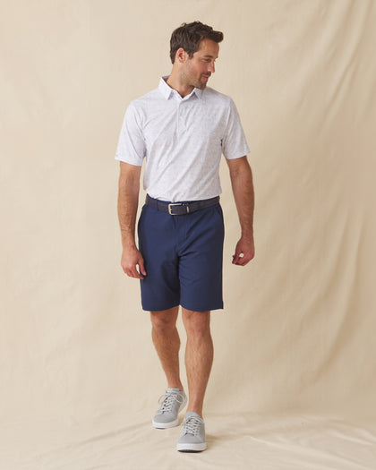 Men's Golden State Polo in Spritz, full length front view on model featuring a white base with an all-over blue and pink California landscape sketch print, paired with navy shorts, a dark leather belt, and gray shoes