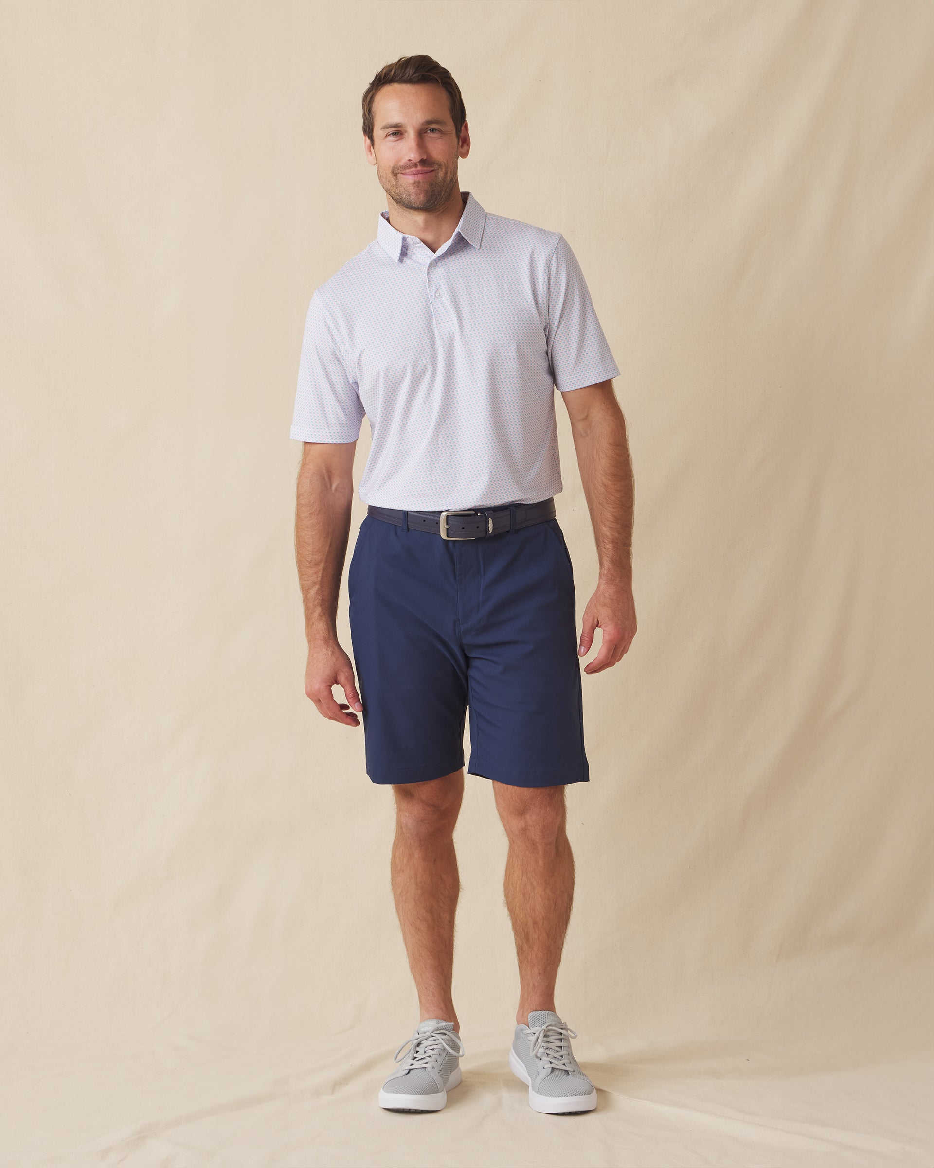 Men's Cocora Polo in Coastal, full length front view on model featuring a white base with an all-over blue and pink palm tree micro print, paired with navy shorts, a dark leather belt, and gray shoes