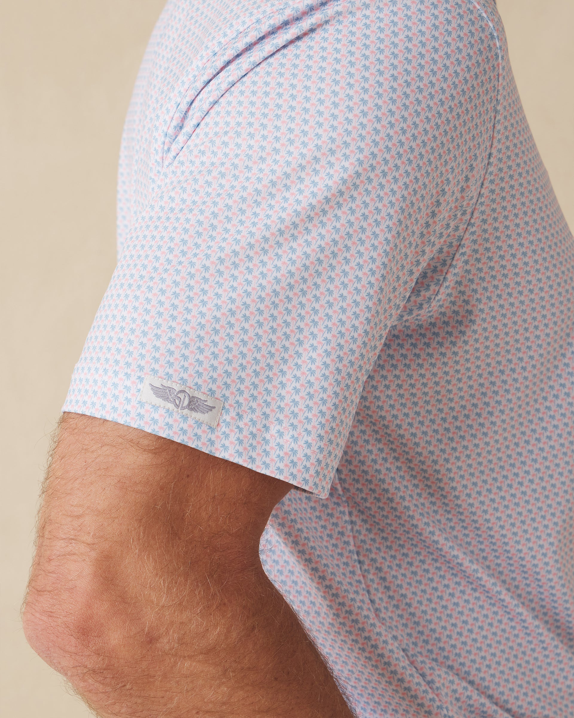 Men's Cocora Polo in Coastal, close-up sleeve view on model highlighting the Straight Down logo patch on the sleeve hem and the all-over blue and pink palm tree micro print on a white base