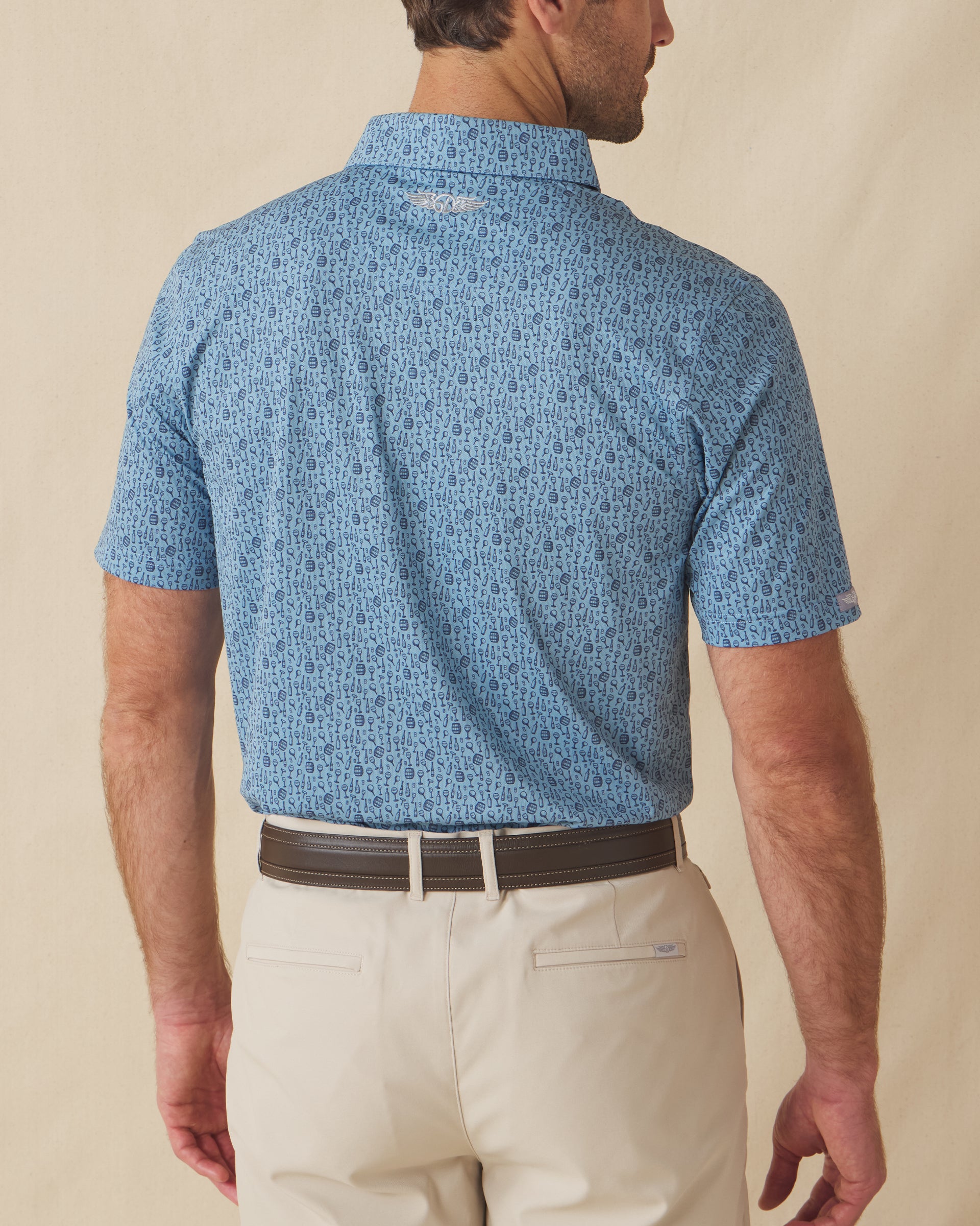 Men's Tannins & Tees Polo in Coastal, back view on model featuring a blue base with a scattered wine bottle, wine glass, barrel, and corkscrew print in navy, with Straight Down logo embroidered at upper back, paired with khaki pants