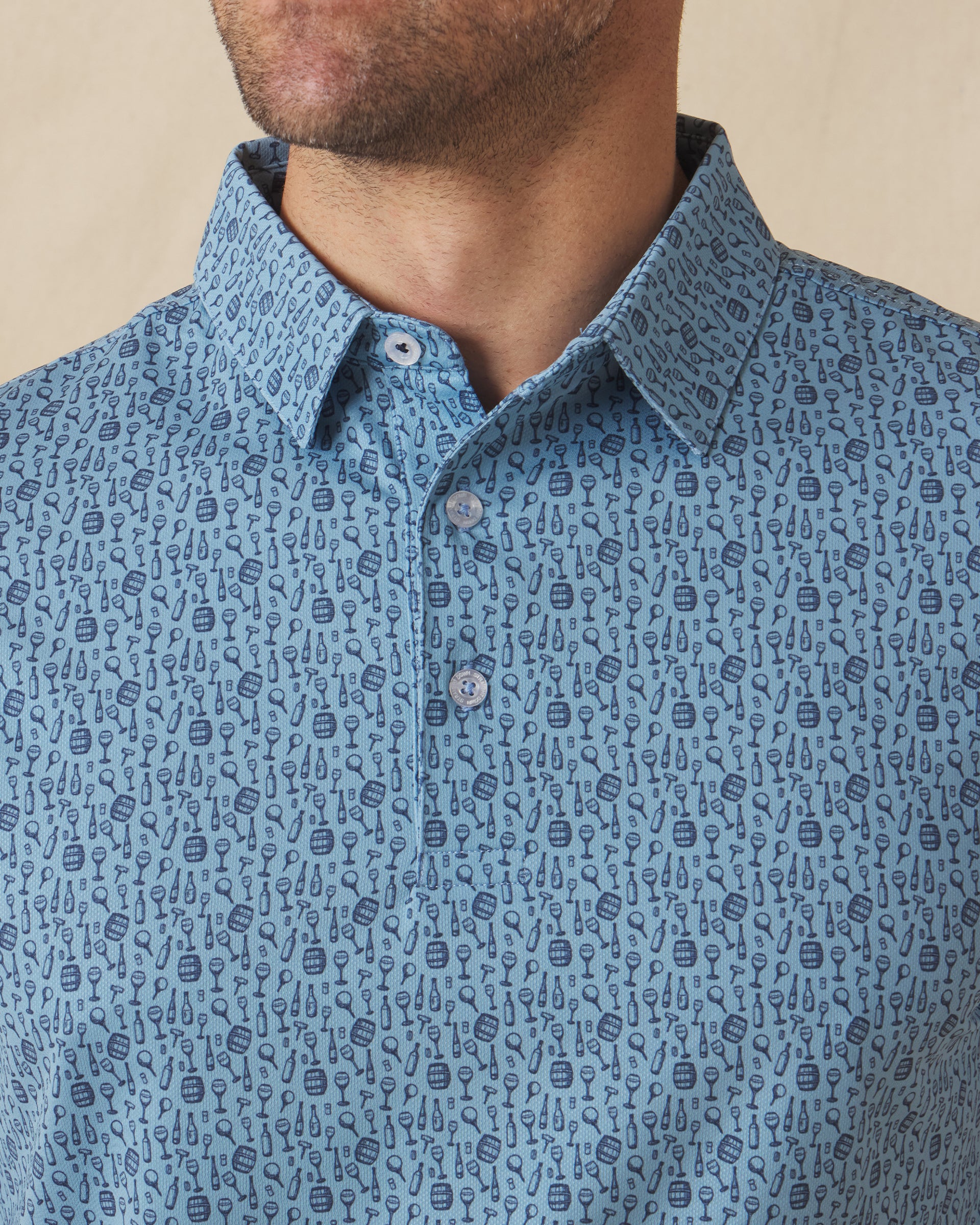 Men's Tannins & Tees Polo in Coastal, close-up front view on model highlighting the scattered wine bottle, wine glass, barrel, and corkscrew print in navy on a blue base