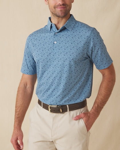 Men's Tannins & Tees Polo in Coastal, front view on model featuring a blue base with a scattered wine bottle, wine glass, barrel, and corkscrew print in navy, paired with khaki pants and a dark leather belt