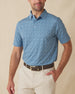 Men's Tannins & Tees Polo in Coastal, front view on model featuring a blue base with a scattered wine bottle, wine glass, barrel, and corkscrew print in navy, paired with khaki pants and a dark leather belt