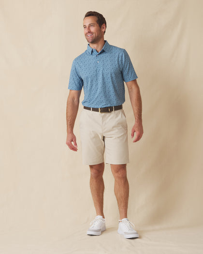 Men's Tannins & Tees Polo in Coastal, full length front view on model featuring a blue base with a scattered wine bottle, wine glass, barrel, and corkscrew print in navy, paired with khaki shorts, a dark leather belt, and white shoes