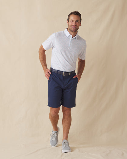 Men's Turtle Island Polo in Coastal, full length model view featuring a white short sleeve polo with an allover sea turtle and coral print in light blue and pink tones, styled with navy shorts and gray shoes