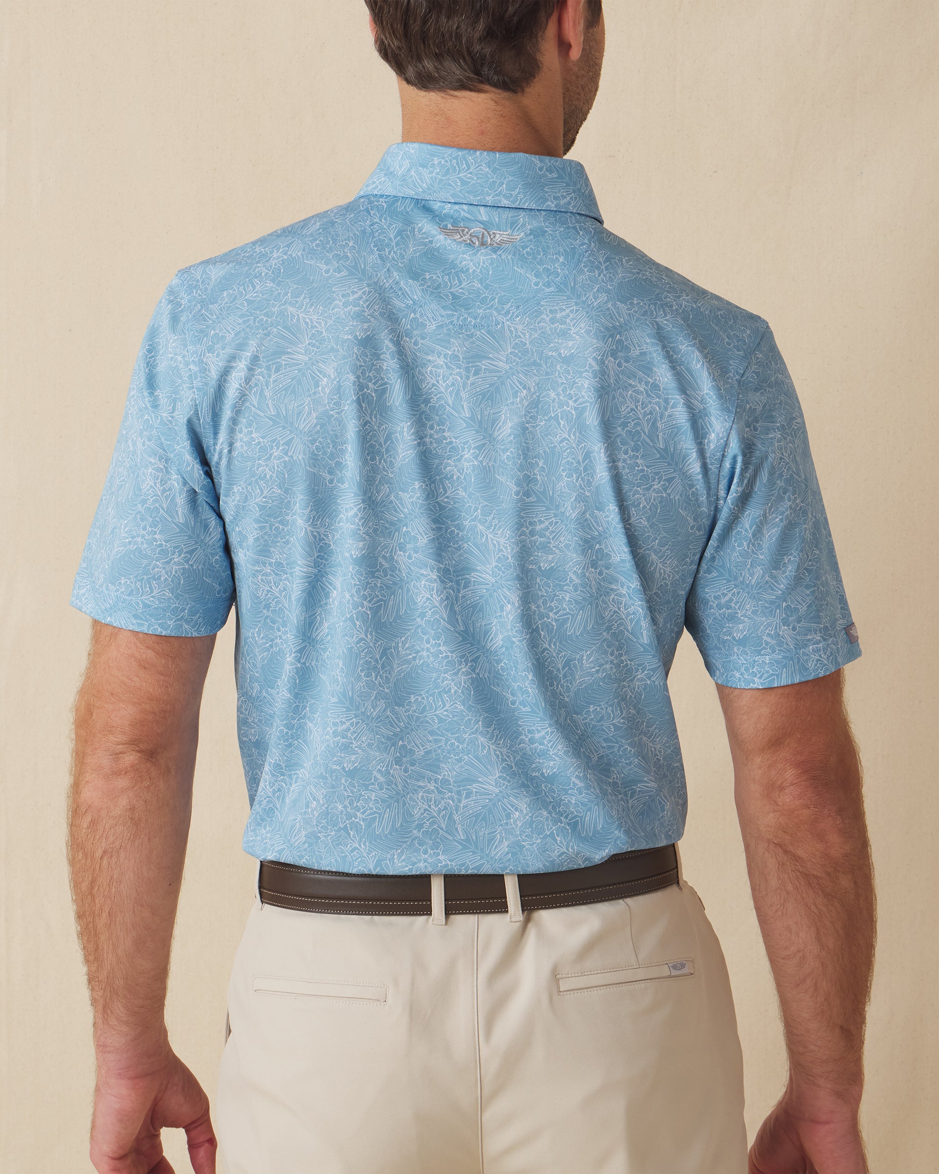 Men's Ka'anapali Polo in Coastal, back model view featuring a medium blue short sleeve polo with an allover white tropical floral and palm leaf line-art print, with an embroidered Straight Down logo at the back collar