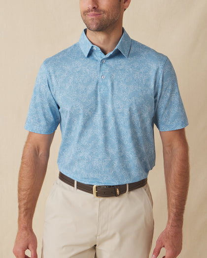 Men's Ka'anapali Polo in Coastal, front model view featuring a medium blue short sleeve polo with an allover white tropical floral and palm leaf line-art print, styled with khaki pants and a leather belt