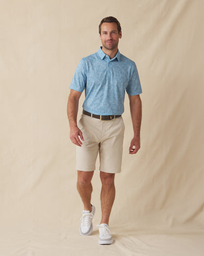 Men's Ka'anapali Polo in Coastal, full length model view featuring a medium blue short sleeve polo with an allover white tropical floral and palm leaf line-art print, styled with khaki shorts and white shoes