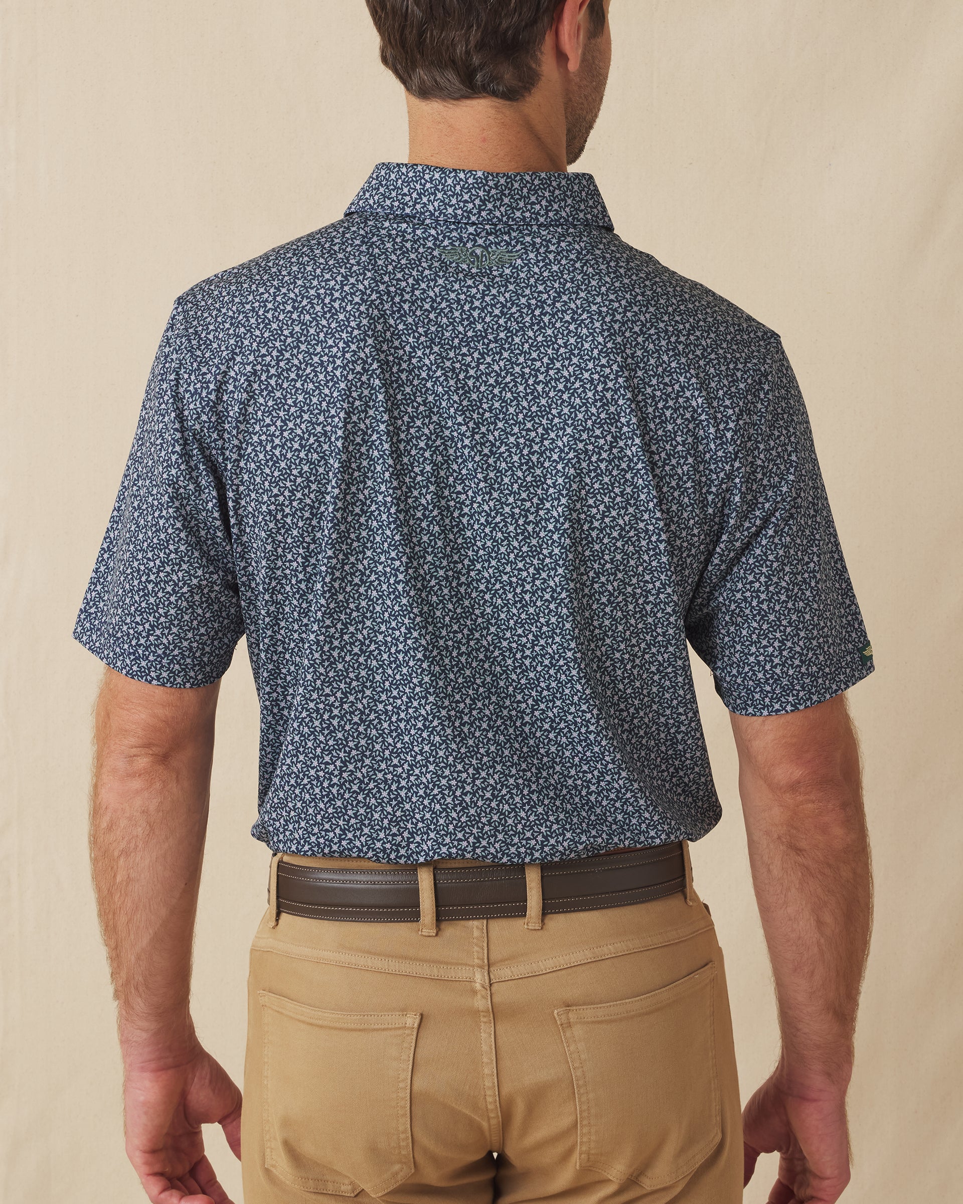 Men's Redbud Polo in New Indigo, back view on model featuring a navy base with a scattered green and light pink floral print and green Straight Down logo embroidered at upper back, paired with khaki pants