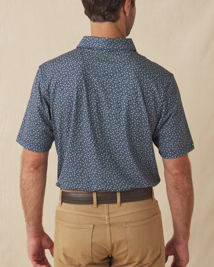 Men's Redbud Polo in New Indigo, back view on model featuring a navy base with a scattered green and light pink floral print and green Straight Down logo embroidered at upper back, paired with khaki pants