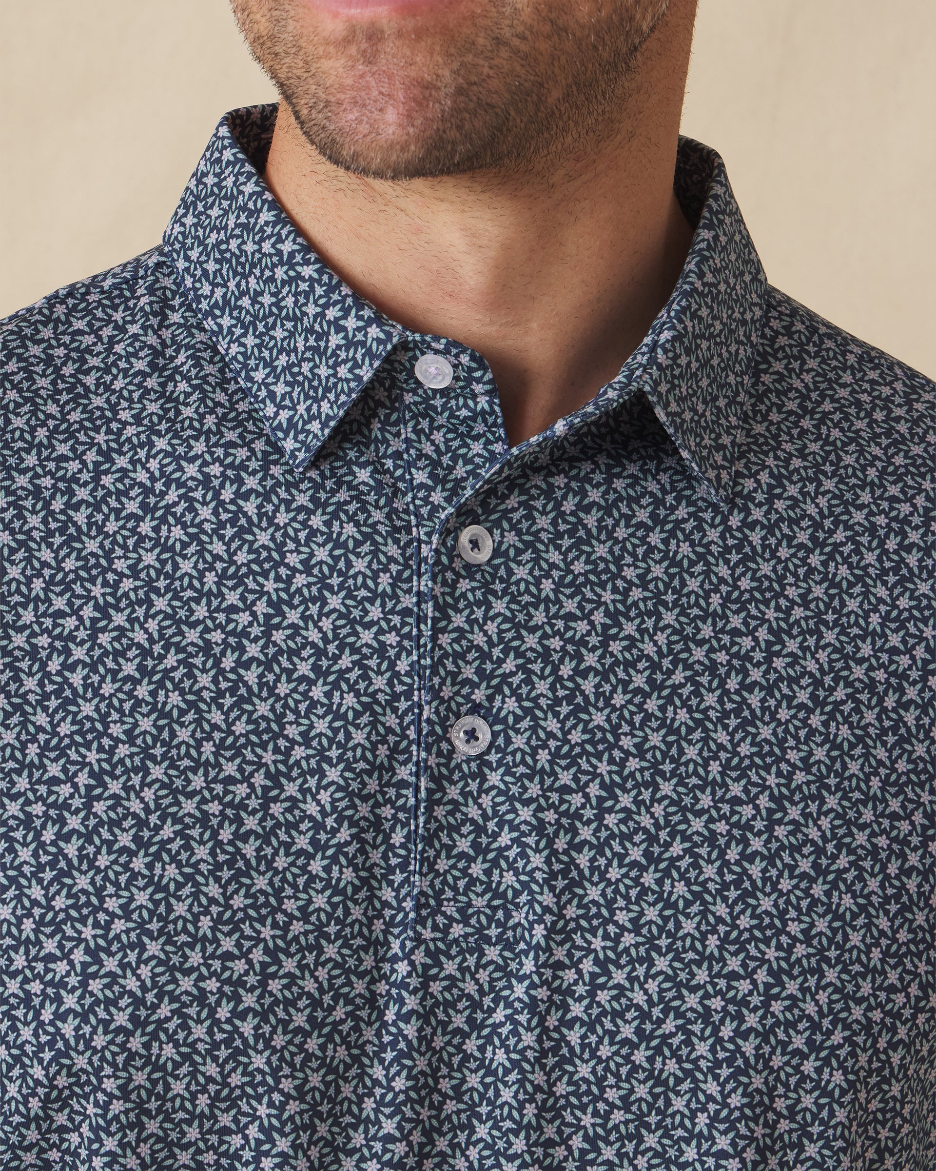 Men's Redbud Polo in New Indigo, close-up detail view on model featuring a navy base with scattered green and light pink floral print and spread collar