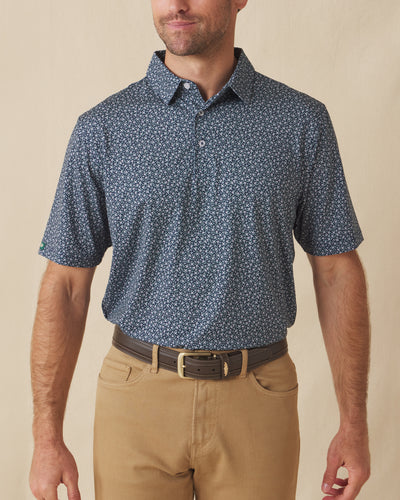 Men's Redbud Polo in New Indigo, front view on model featuring a navy base with a scattered green and light pink floral print, paired with khaki pants and a leather belt