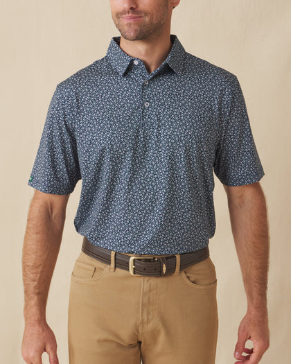 Men's Redbud Polo in New Indigo, front view on model featuring a navy base with a scattered green and light pink floral print, paired with khaki pants and a leather belt