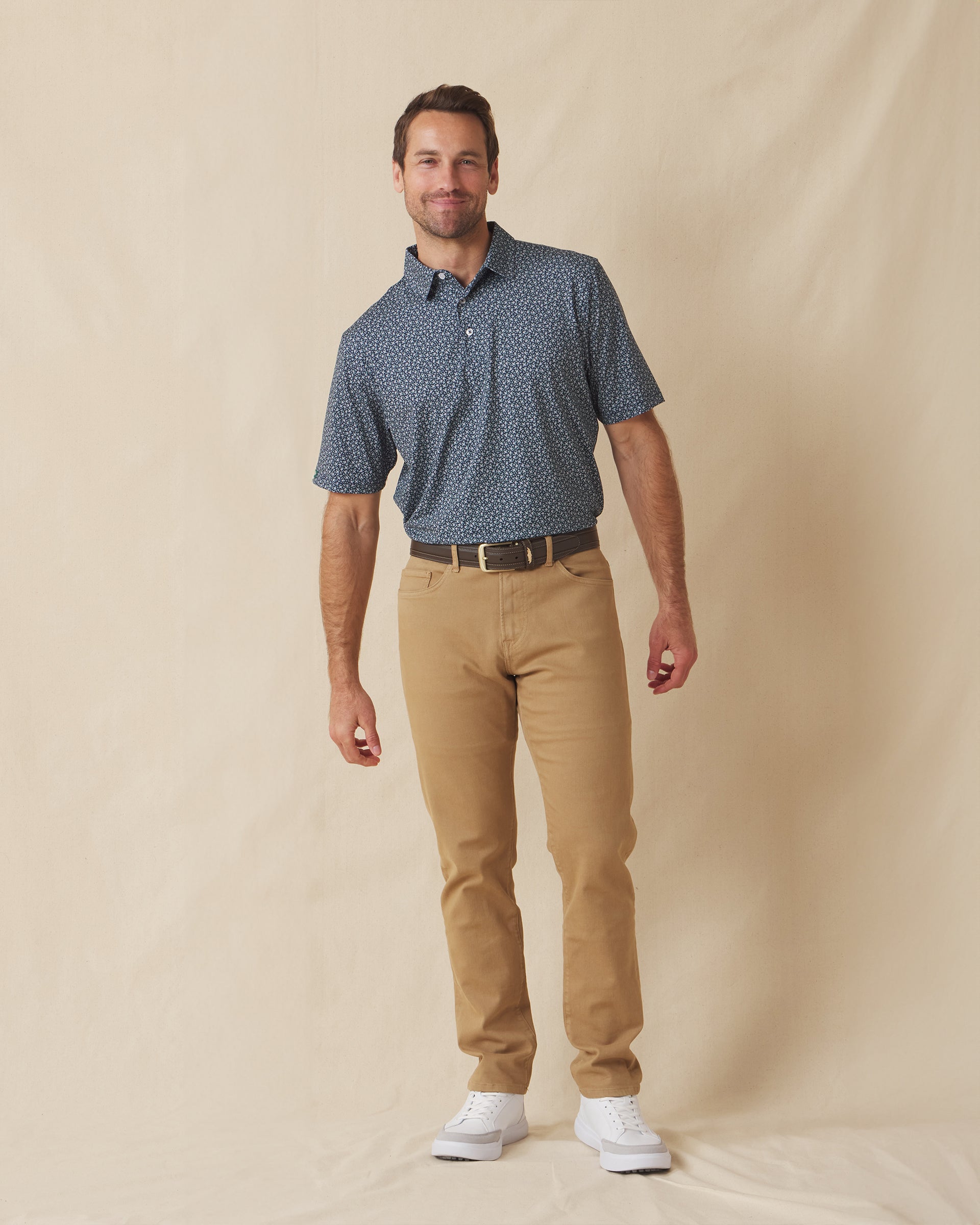 Men's Redbud Polo in New Indigo, full-length front view on model featuring a navy base with a scattered green and light pink floral print, paired with khaki pants and white golf shoes