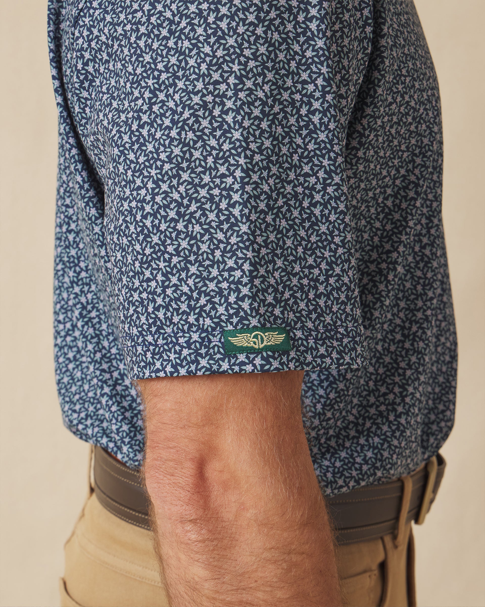 Men's Redbud Polo in New Indigo, close-up detail view on model featuring green and gold Straight Down logo sleeve tab on navy base with scattered green and light pink floral print