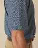 Men's Redbud Polo in New Indigo, close-up detail view on model featuring green and gold Straight Down logo sleeve tab on navy base with scattered green and light pink floral print