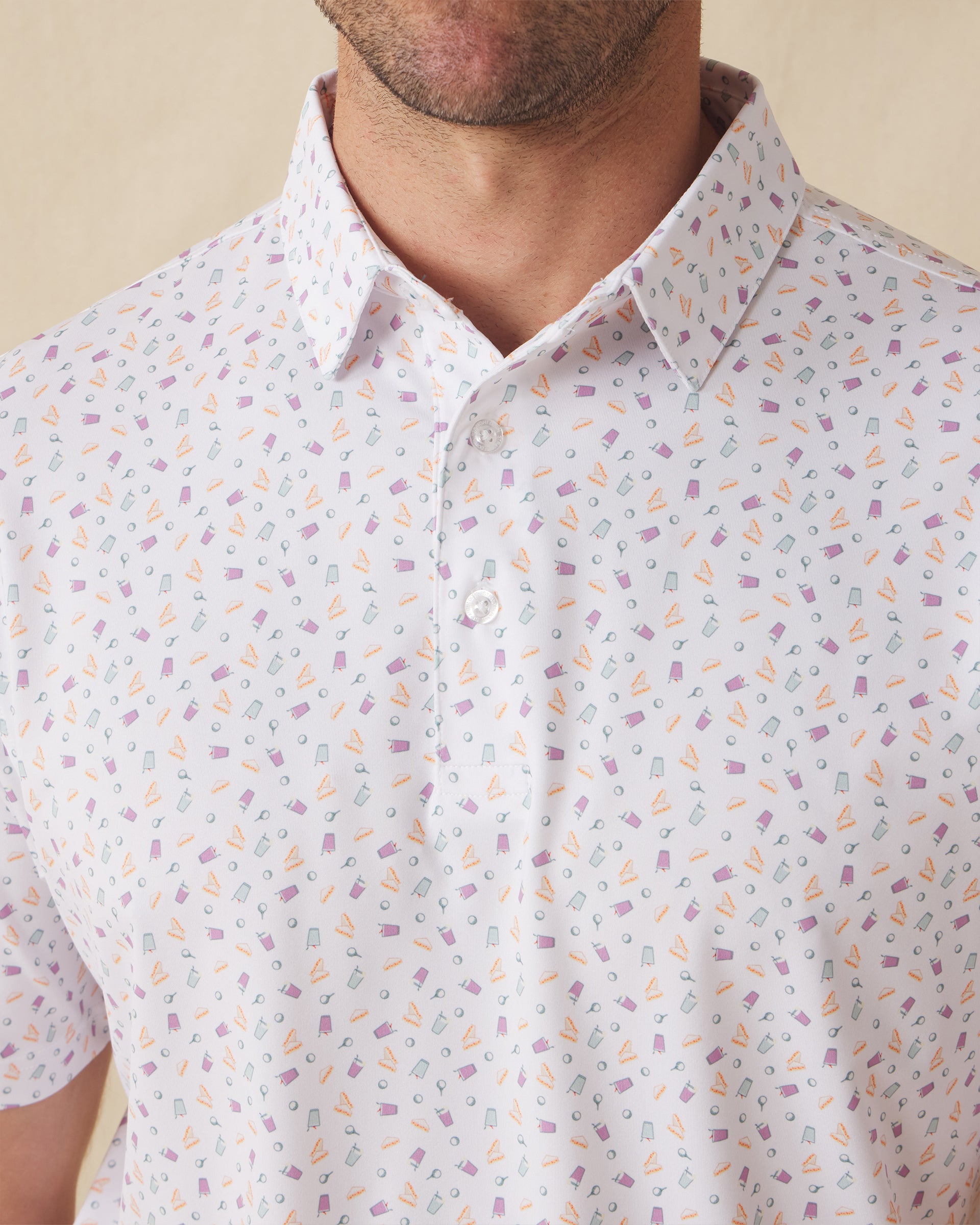 Men's Pimento Cheese Polo in White, close-up front view on model highlighting a scattered golf, sandwich, and drink print in purple, teal, and orange on a white base