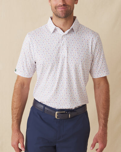 Men's Pimento Cheese Polo in White, front view on model featuring a white base with a scattered golf, sandwich, and drink print in purple, teal, and orange, paired with navy pants and a dark leather belt