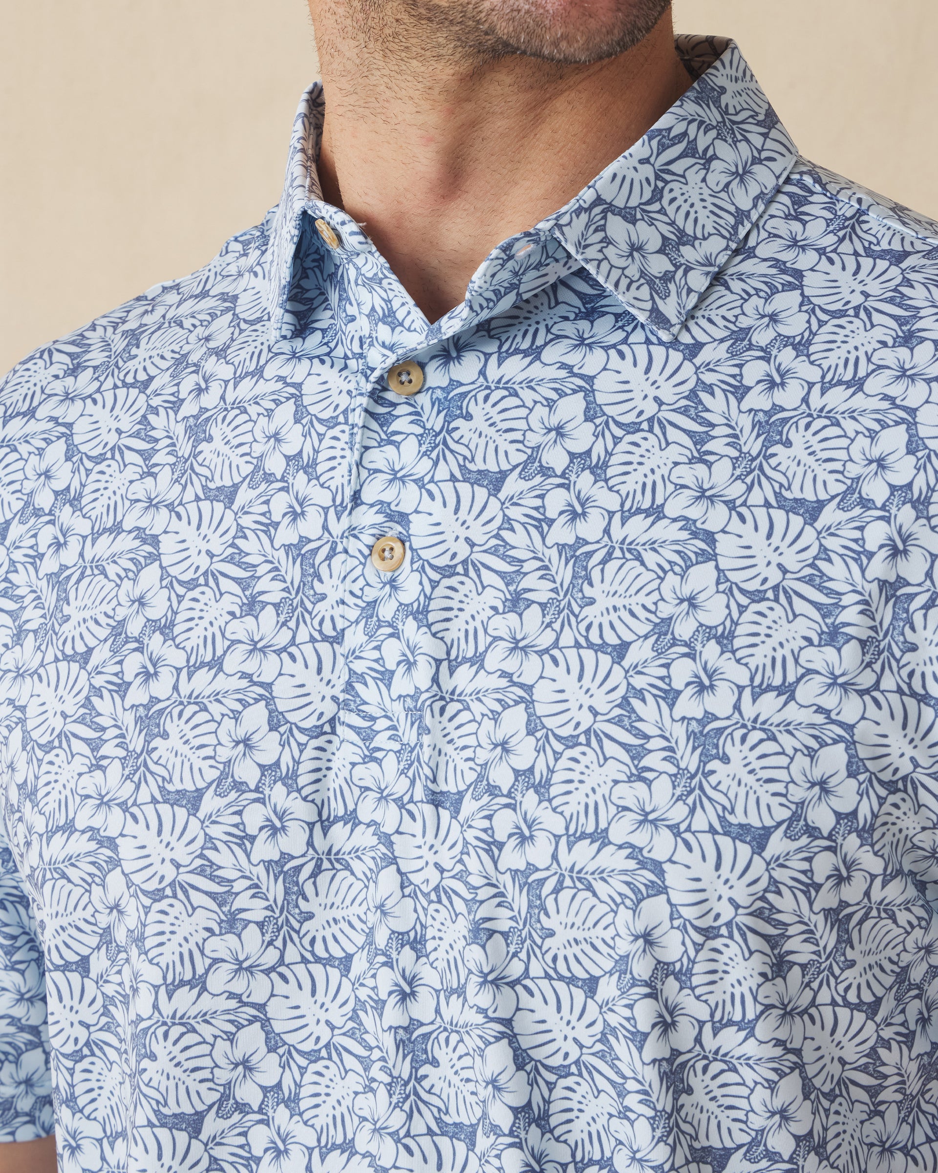 Men's Lihu'e Polo in Sea Spray, collar detail view featuring an allover navy hibiscus flower and monstera leaf print on a light blue performance fabric with natural wood-tone buttons