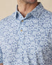Men's Lihu'e Polo in Sea Spray, collar detail view featuring an allover navy hibiscus flower and monstera leaf print on a light blue performance fabric with natural wood-tone buttons