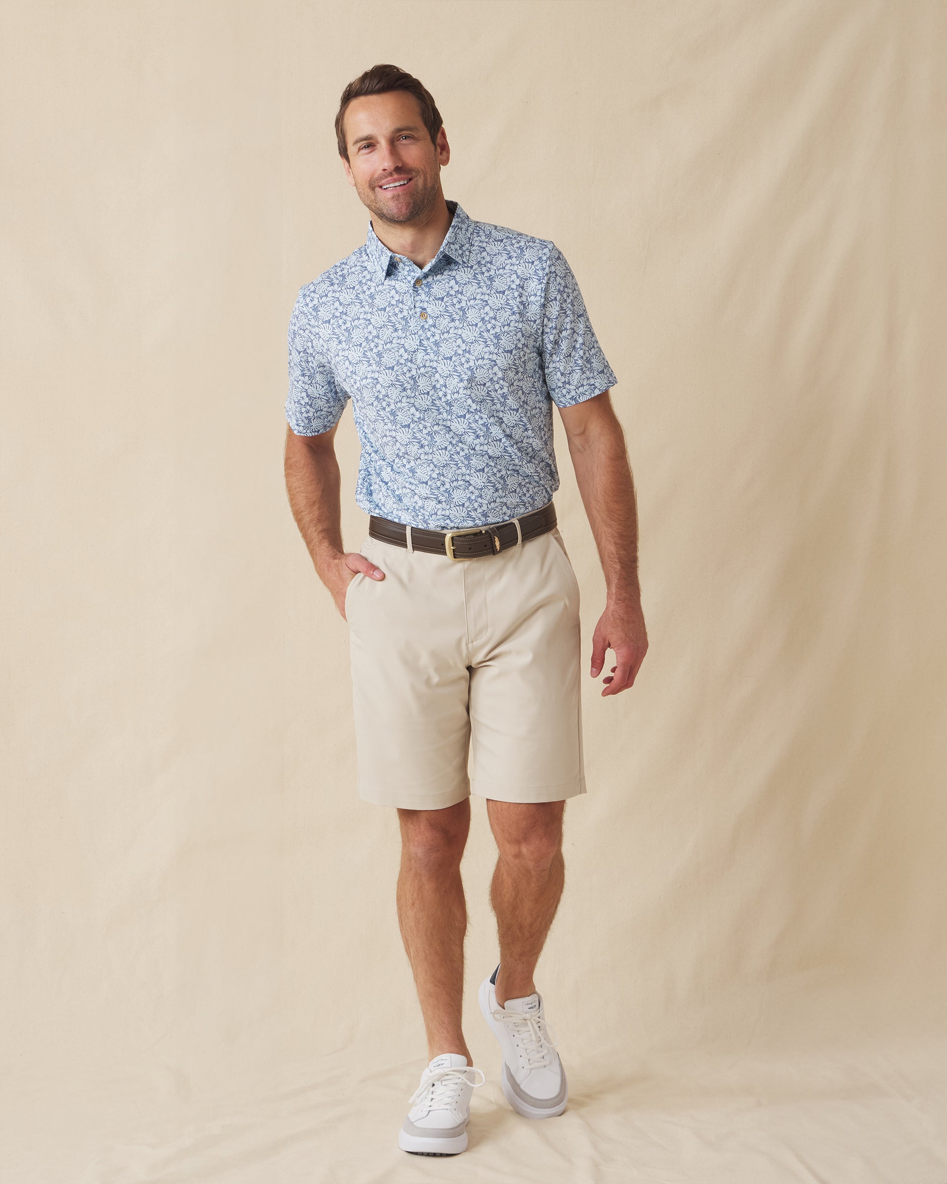 Men's Lihu'e Polo in Sea Spray, full length model view featuring a light blue short sleeve polo with an allover navy hibiscus flower and monstera leaf print, styled with khaki shorts and white shoes