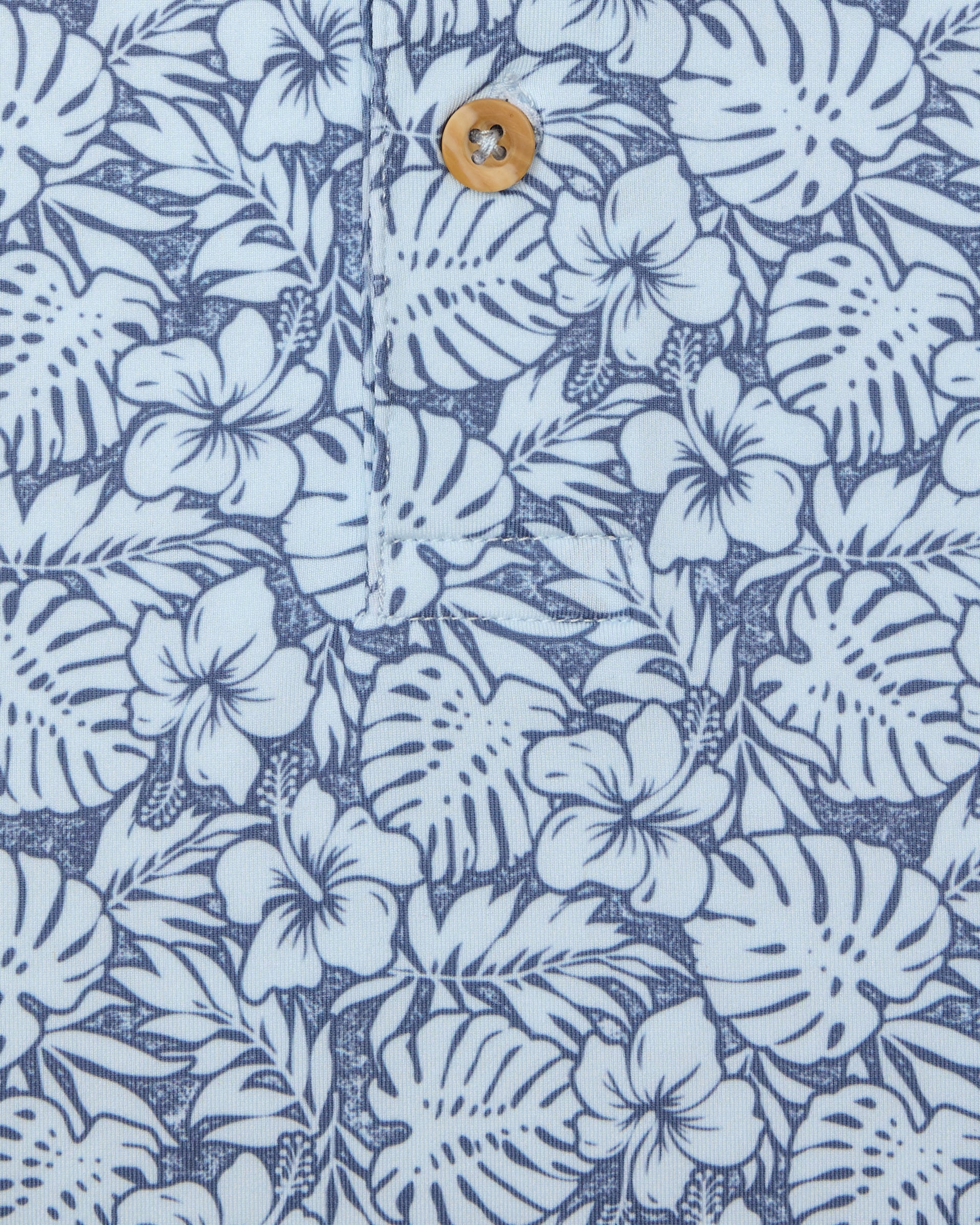 Men's Lihu'e Polo in Sea Spray, close-up fabric detail view featuring an allover navy hibiscus flower and monstera leaf print on a light blue performance fabric with a natural wood-tone button