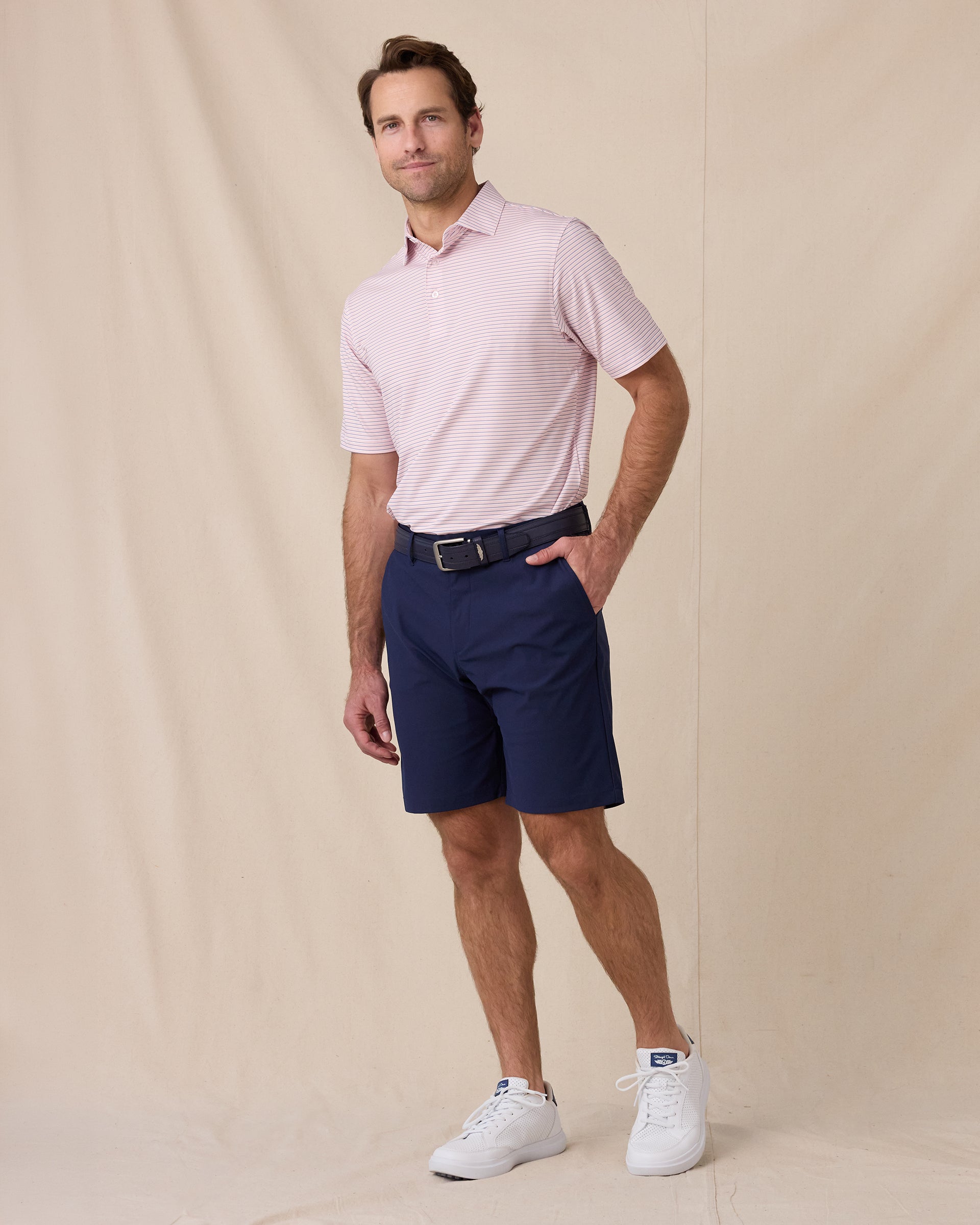 Full-length view of a male model wearing the Channel Stripe Polo in Seashell with navy shorts, a navy belt, and white shoes.