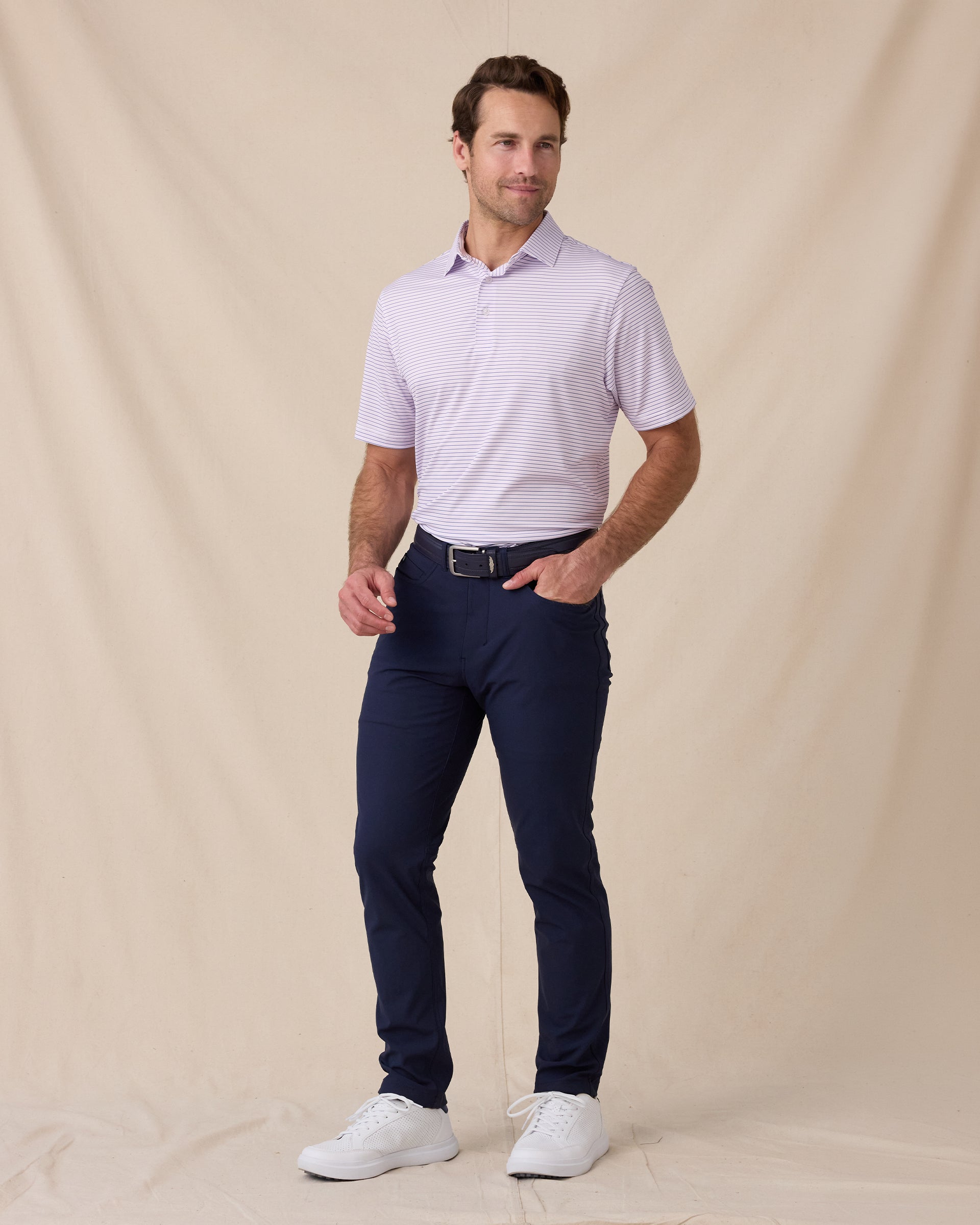 Model wearing the Men's Channel Stripe Golf Polo in Lilac/Tidal with navy golf pants and white golf shoes.