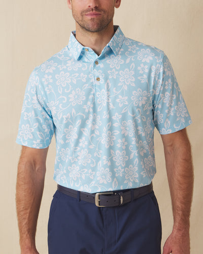Men's Napali Coast Polo in Gemstone, front model view featuring a light aqua blue short sleeve polo with an allover white Hawaiian floral and vine print, styled with navy pants and a leather belt